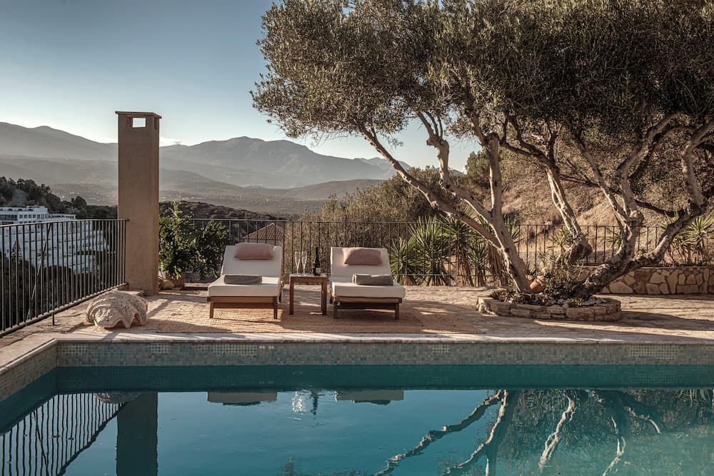 A serene poolside terrace with lounge chairs, a stone chimney, and lush olive trees offers breathtaking mountain views, ideal for a tranquil luxury retreat.