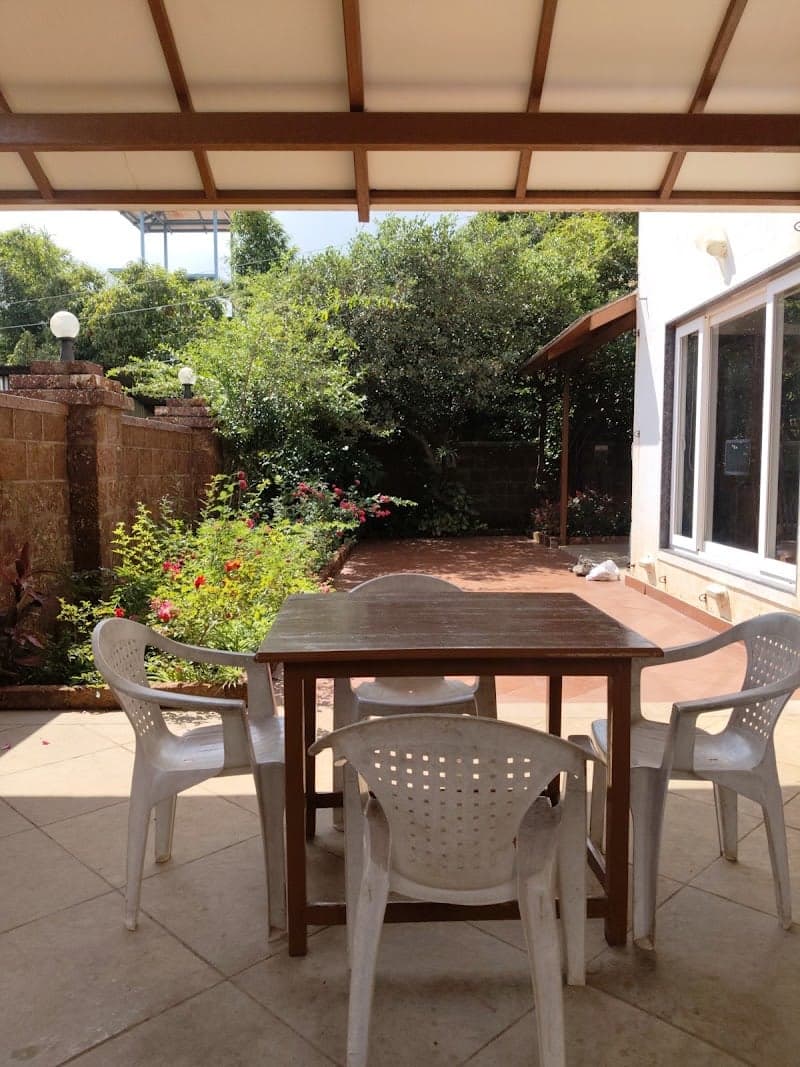 A serene outdoor patio at Anupama Bunglow in Mahabaleshwar features a wooden table with white plastic chairs under a shaded roof, surrounded by lush greenery, ideal for travelers seeking a peaceful retreat.