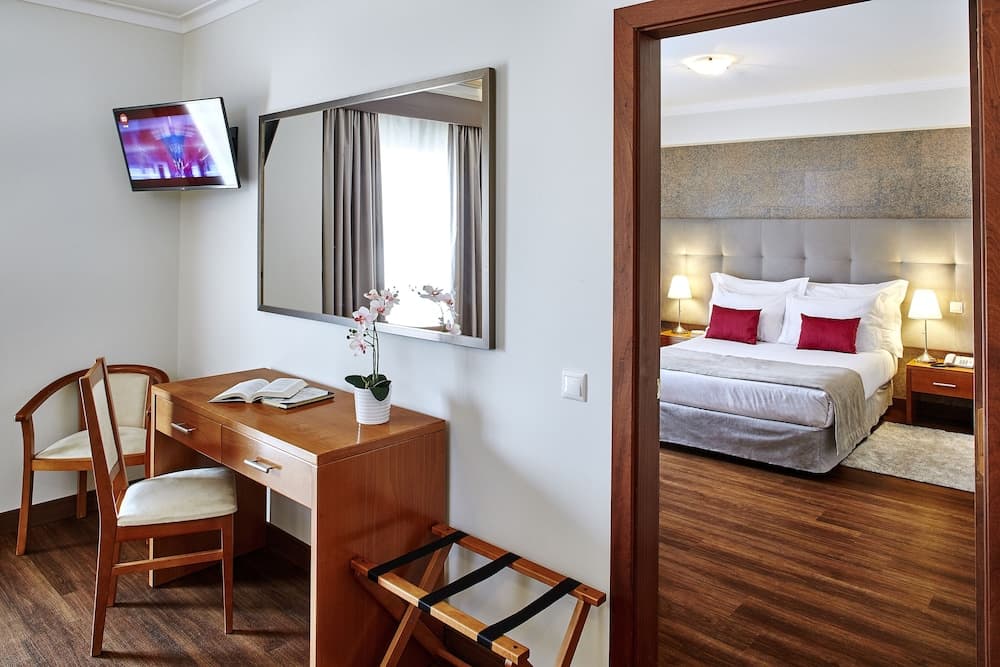 A cozy hotel room features a wooden desk with chairs, a flat-screen TV, a mirror reflecting soft light, and a neatly made bed with red accents, ideal for travelers seeking comfort and functionality.