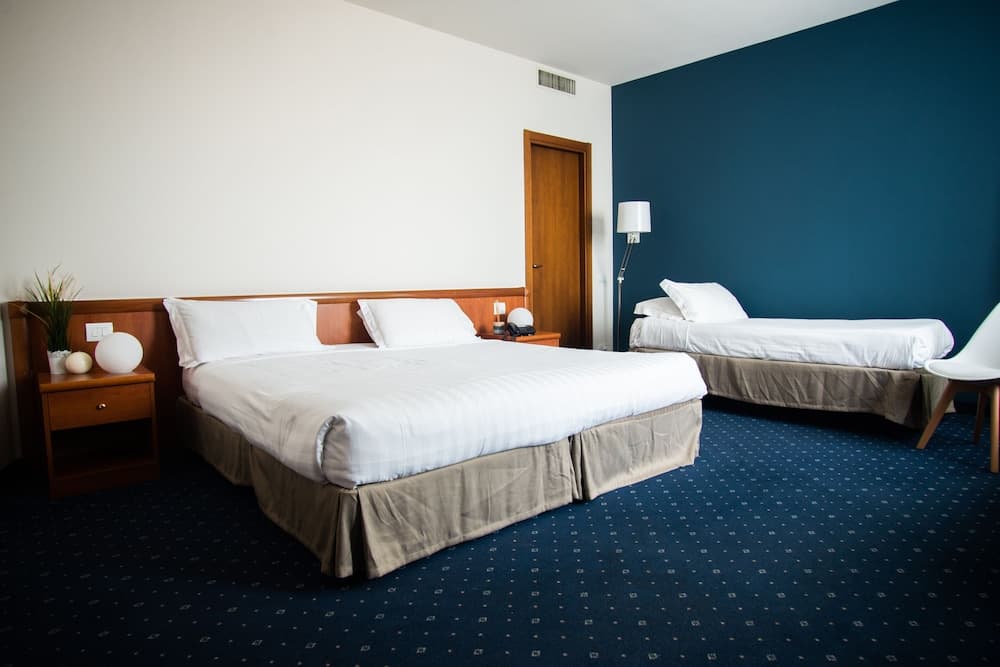 A cozy hotel room features a neatly made double bed and a single bed against a calming blue wall, complemented by wooden furniture and a modern lamp, ideal for travelers seeking comfort and simplicity.