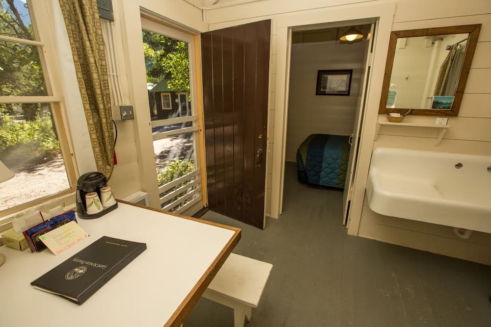 A cozy cabin interior features a small dining table with coffee supplies, a bathroom sink, and an open door leading to a serene outdoor view, ideal for travelers seeking a rustic retreat.