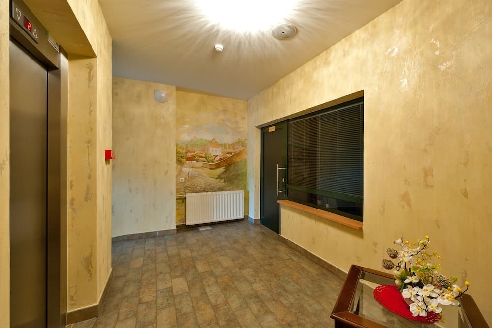 A warmly lit hotel lobby features textured beige walls, a scenic mural, an elevator, a window with blinds, and a floral arrangement on a table, creating a cozy and inviting atmosphere for travelers seeking comfort.