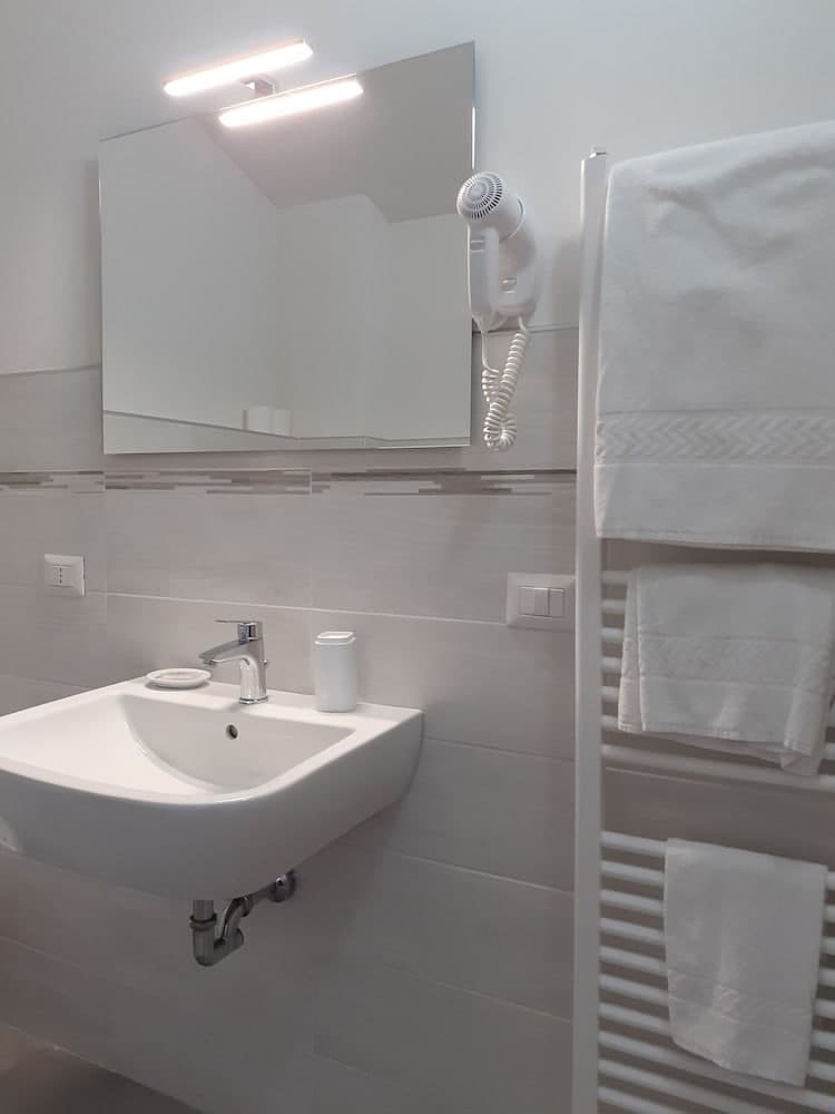 A modern, minimalist bathroom features a sleek sink, a wall-mounted hairdryer, neatly folded towels on a radiator, and a large mirror illuminated by soft lighting, ideal for travelers seeking clean and functional accommodations.