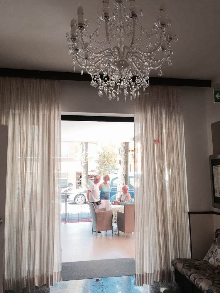 A cozy indoor lounge with elegant curtains and a sparkling chandelier frames a serene outdoor patio where travelers can relax under the shade of trees, ideal for leisurely getaways.