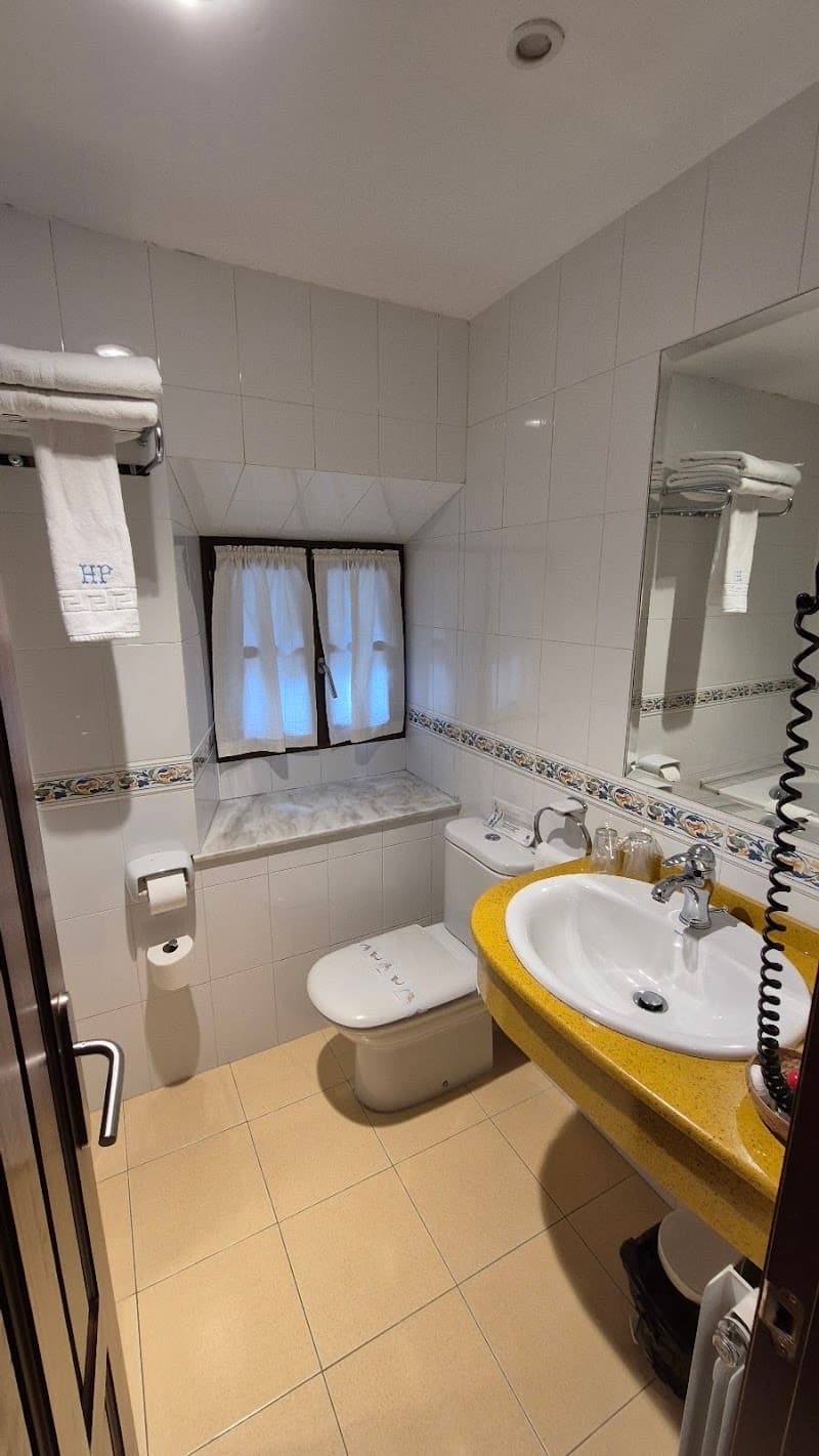 A cozy hotel bathroom in Asturias, Spain, features white tiled walls, a yellow countertop sink, a bidet, and a window with curtains, creating a clean and inviting atmosphere ideal for travelers seeking comfort during their stay.