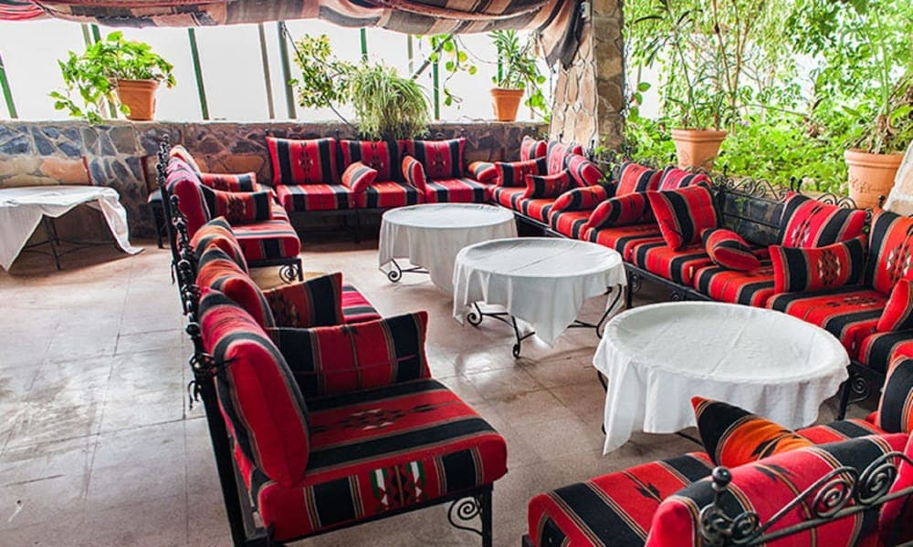 A cozy indoor lounge with vibrant red and black patterned sofas, white tableclothed tables, and lush greenery creates an inviting atmosphere perfect for travelers seeking relaxation in a cultural retreat setting.