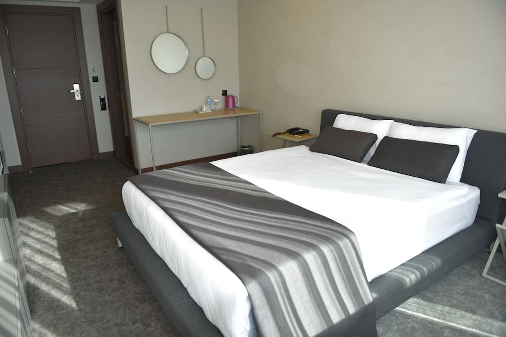 A cozy hotel room features a neatly made bed with gray and white linens, a small desk with mirrors above it, and soft lighting, ideal for travelers seeking comfort and relaxation.
