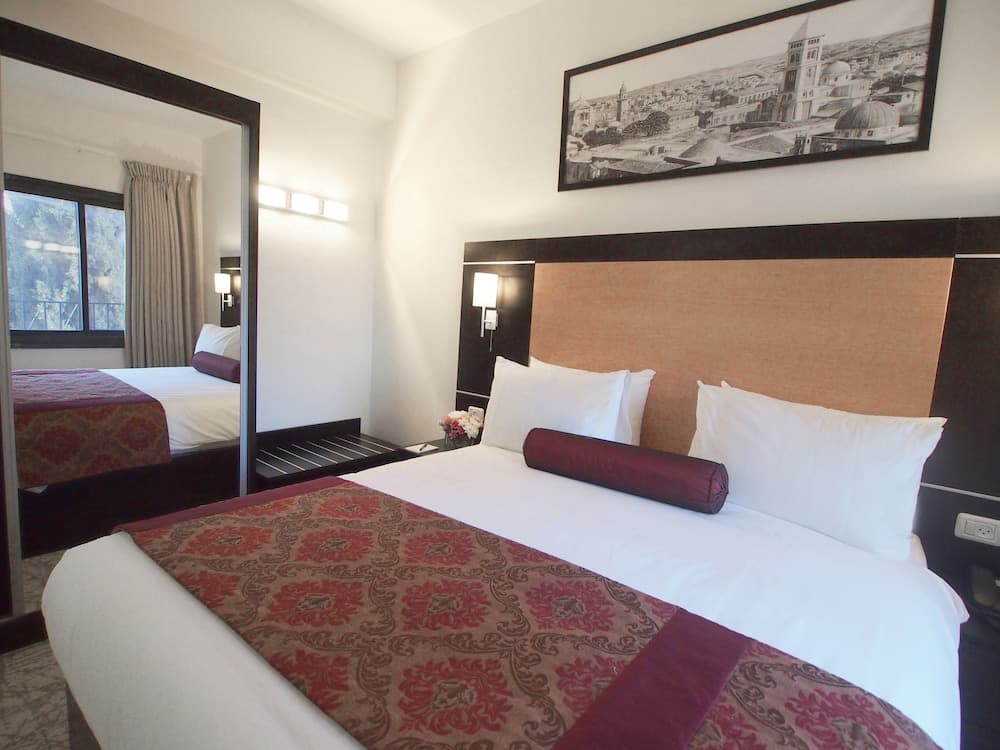 A cozy hotel room features a neatly made bed with a patterned throw, a large mirror reflecting the window view, and a wall-mounted cityscape artwork, ideal for travelers seeking comfort and a touch of local charm.