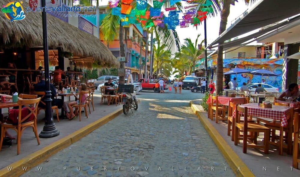 A vibrant cobblestone street lined with colorful papel picado banners, outdoor dining tables, and palm trees creates a lively and inviting atmosphere in a bustling tropical destination perfect for leisurely strolls and cultural exploration.