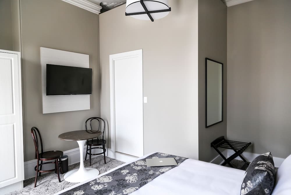 A cozy hotel room features neutral tones, a neatly made bed with patterned accents, a small dining table with chairs, a wall-mounted TV, and a modern light fixture, ideal for travelers seeking comfort and simplicity.
