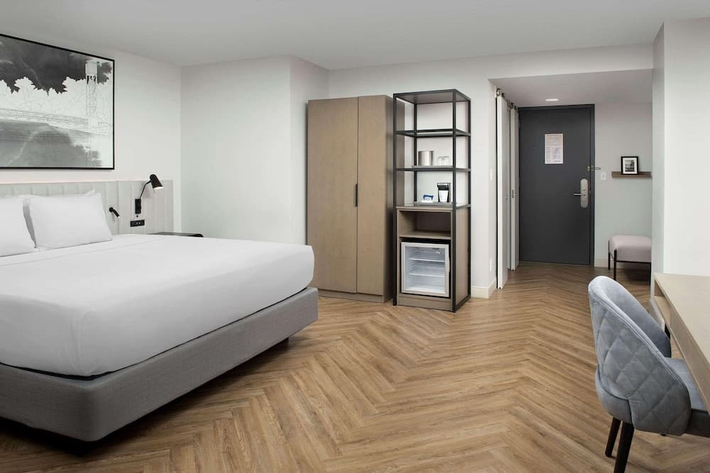 A modern hotel room features a cozy bed, sleek wooden flooring, a mini-fridge, and a minimalist desk area, creating a serene and functional space ideal for travelers seeking comfort and convenience.
