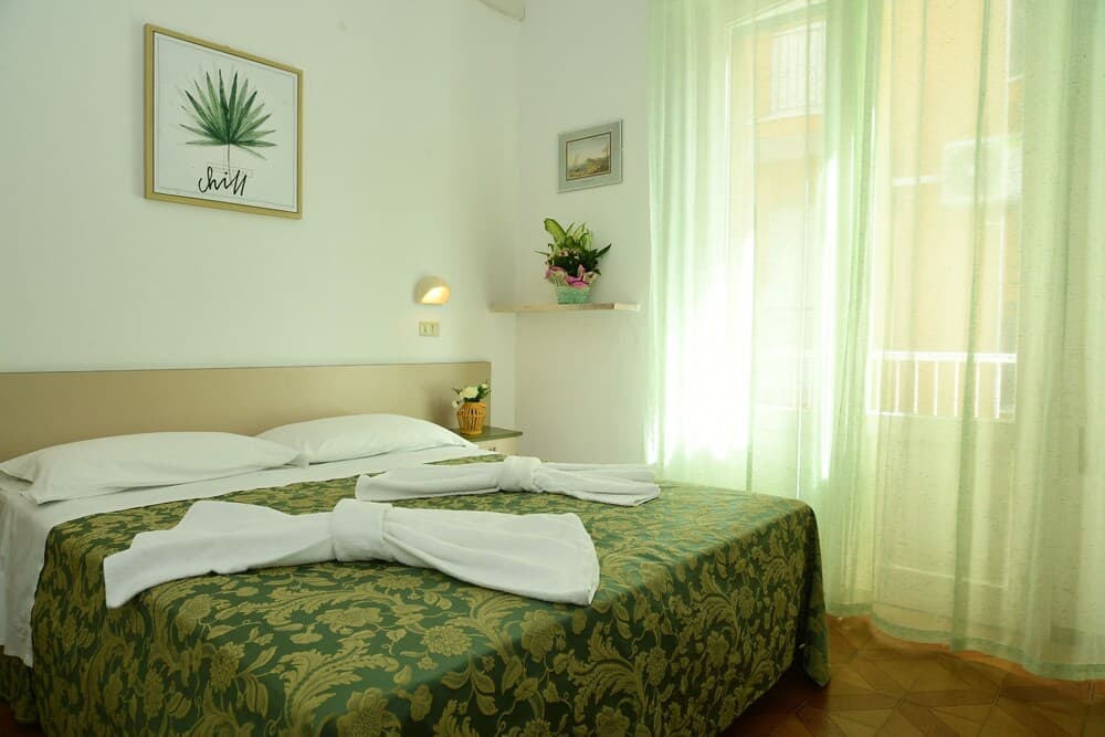 A cozy hotel room with a neatly made bed adorned with green floral bedding and towel swans, soft natural light filtering through sheer curtains, and decorative wall art creates a serene atmosphere ideal for travelers seeking relaxation.