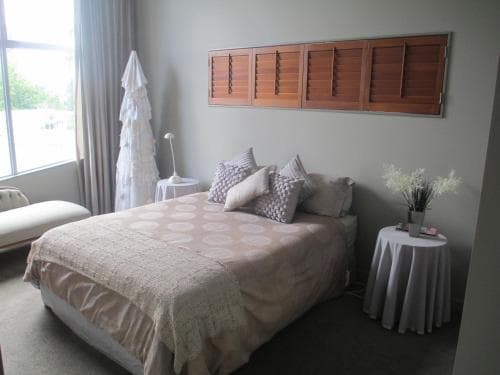 A cozy bedroom with a neatly made bed adorned with patterned pillows, wooden shutters above, and soft natural light filtering through sheer curtains, creating a serene atmosphere ideal for a relaxing getaway.