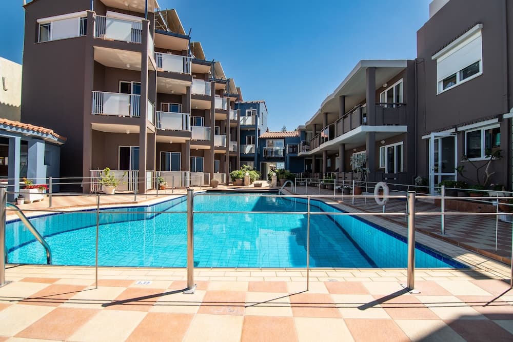 A serene resort scene features a sparkling blue pool surrounded by modern apartment buildings under a clear sky, ideal for vacationers seeking relaxation and comfort.