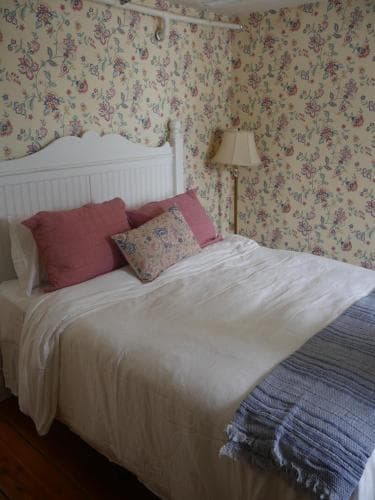 A cozy bedroom with floral wallpaper exudes a vintage charm, featuring a neatly made bed adorned with pink pillows and a blue blanket, ideal for travelers seeking a quaint and comfortable stay.