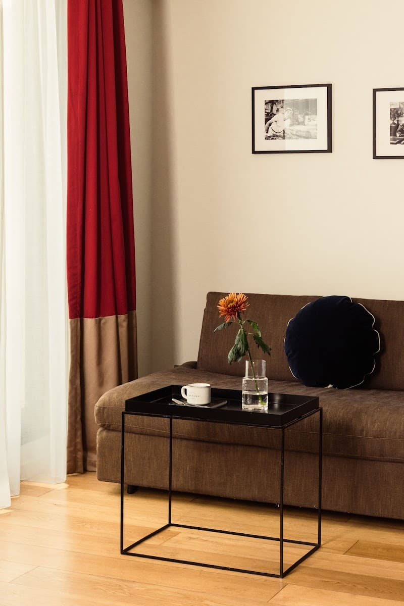 A cozy corner in a Parisian hotel room features a brown sofa adorned with a navy blue pillow, a modern black coffee table holding a vase with an orange flower and a mug, set against a backdrop of red and beige curtains and framed black-and-white artwork, ideal for travelers seeking a stylish and comfortable stay.