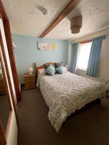 A cozy bedroom with a neatly made bed adorned in floral bedding, blue curtains framing a bright window, wooden beams on the ceiling, and a serene atmosphere ideal for a relaxing getaway.