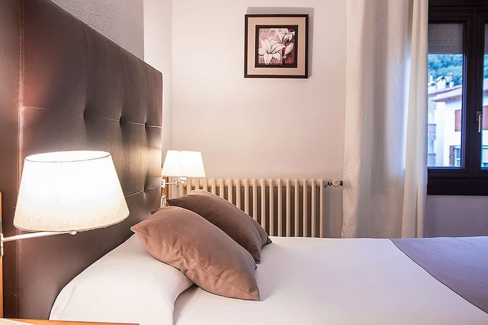 A cozy hotel room features a neatly made bed with brown pillows, soft lighting from bedside lamps, a floral artwork on the wall, and a window view of buildings and greenery, ideal for travelers seeking comfort and relaxation.