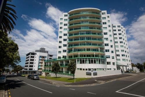 A modern, curved high-rise building stands prominently on a sunny day in an urban area, featuring balconies and greenery, ideal for travelers seeking upscale accommodation with city views.