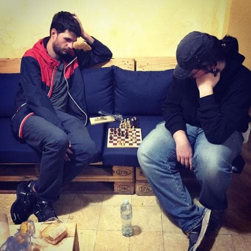 Two individuals deeply engrossed in a chess game sit on a cozy blue couch in a rustic café setting, surrounded by casual attire and a water bottle, evoking a relaxed yet competitive atmosphere ideal for travelers seeking a quiet retreat or intellectual engagement.