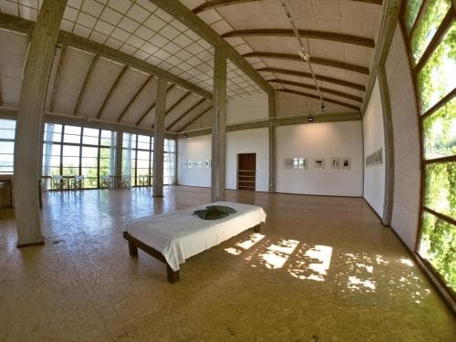 A serene art gallery space features a minimalist bed under a vaulted wooden ceiling, surrounded by natural light and greenery, ideal for travelers seeking peaceful retreats and cultural experiences.