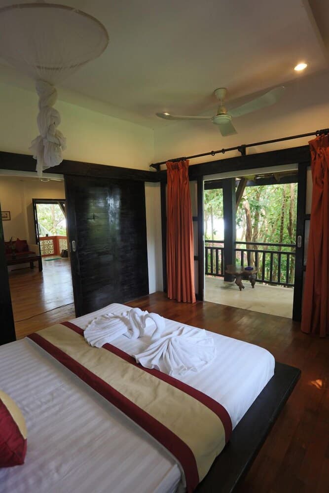 A cozy tropical villa bedroom with wooden floors, vibrant red curtains, and a balcony overlooking lush greenery, ideal for travelers seeking relaxation and nature immersion.