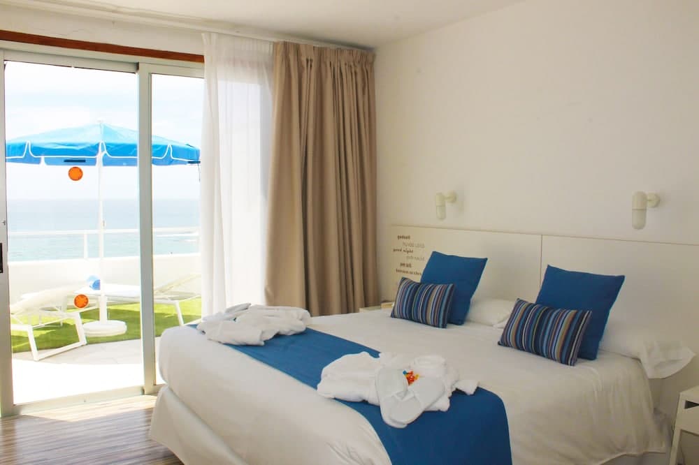 A serene hotel room with a cozy bed adorned in blue and white linens opens to a balcony overlooking the ocean under a bright blue umbrella, ideal for travelers seeking relaxation by the sea.