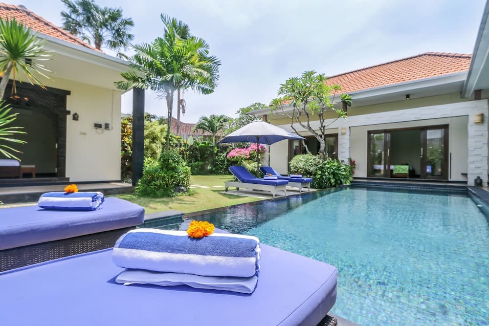 A serene tropical villa poolside scene features plush lounge chairs adorned with neatly folded towels and vibrant orange flowers, surrounded by lush greenery and traditional architecture, ideal for a relaxing vacation getaway.