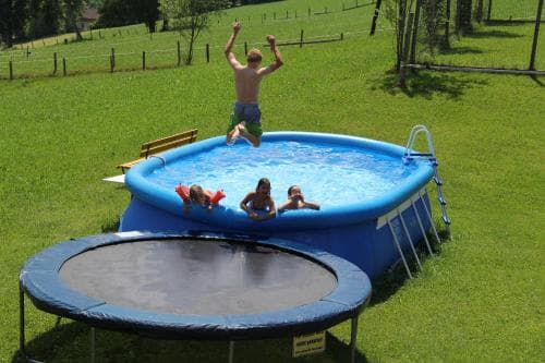A joyful family scene unfolds in a lush green backyard where children play in an above-ground pool and on a trampoline, ideal for summer vacation activities and family bonding.