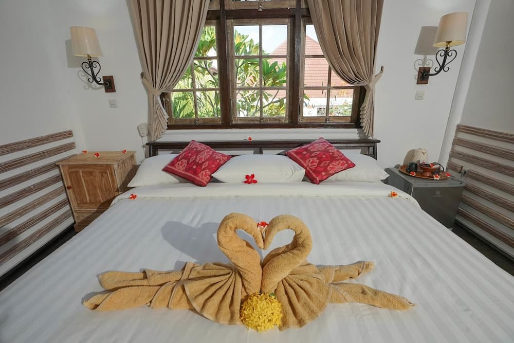 A cozy hotel bedroom with a neatly arranged bed featuring towel swans in a heart shape, red floral accents, wooden furnishings, and a window view of lush greenery, ideal for romantic getaways.
