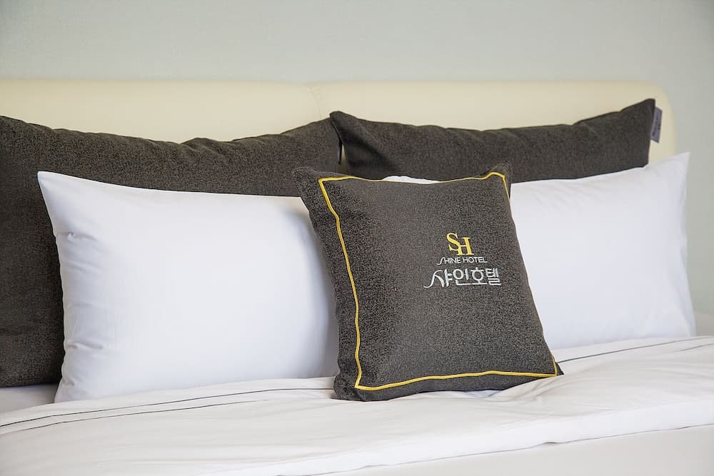 A cozy hotel bed adorned with plush pillows featuring the "Shine Hotel" logo creates a welcoming atmosphere ideal for travelers seeking comfort and relaxation.