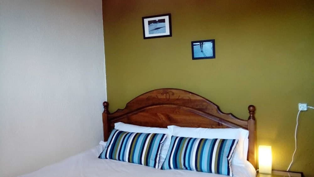 A cozy bedroom with a wooden bed adorned with striped pillows, framed artwork on a mustard-yellow wall, and a warm bedside lamp creates an inviting atmosphere ideal for travelers seeking comfort and relaxation.