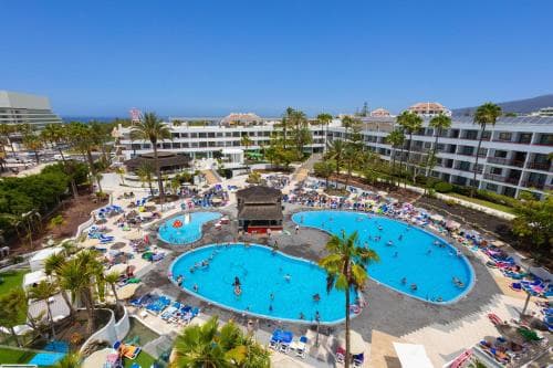 A vibrant resort scene featuring multiple sparkling blue pools surrounded by lounging guests under palm trees, ideal for tropical vacation inspiration.