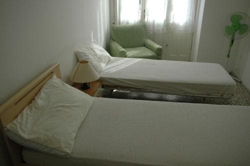 A cozy hotel room features two neatly made beds, a green armchair by the window, and a fan, creating a simple and inviting atmosphere ideal for travelers seeking restful accommodations.