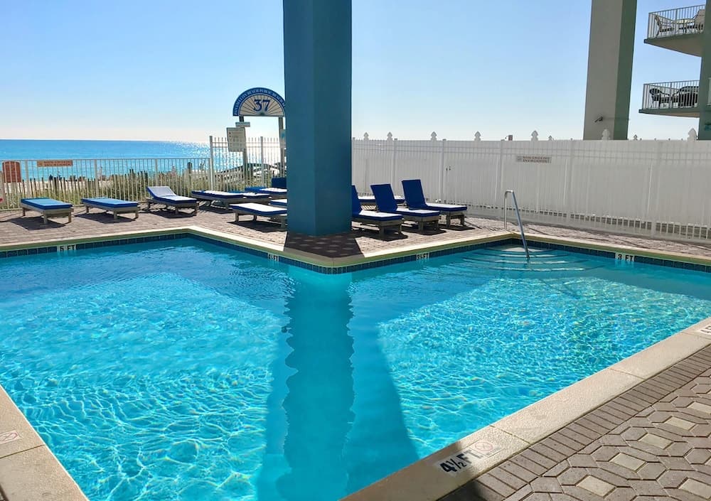 A serene beachside resort pool area features crystal-clear water, blue lounge chairs, and a view of the ocean, ideal for vacation relaxation and family getaways.