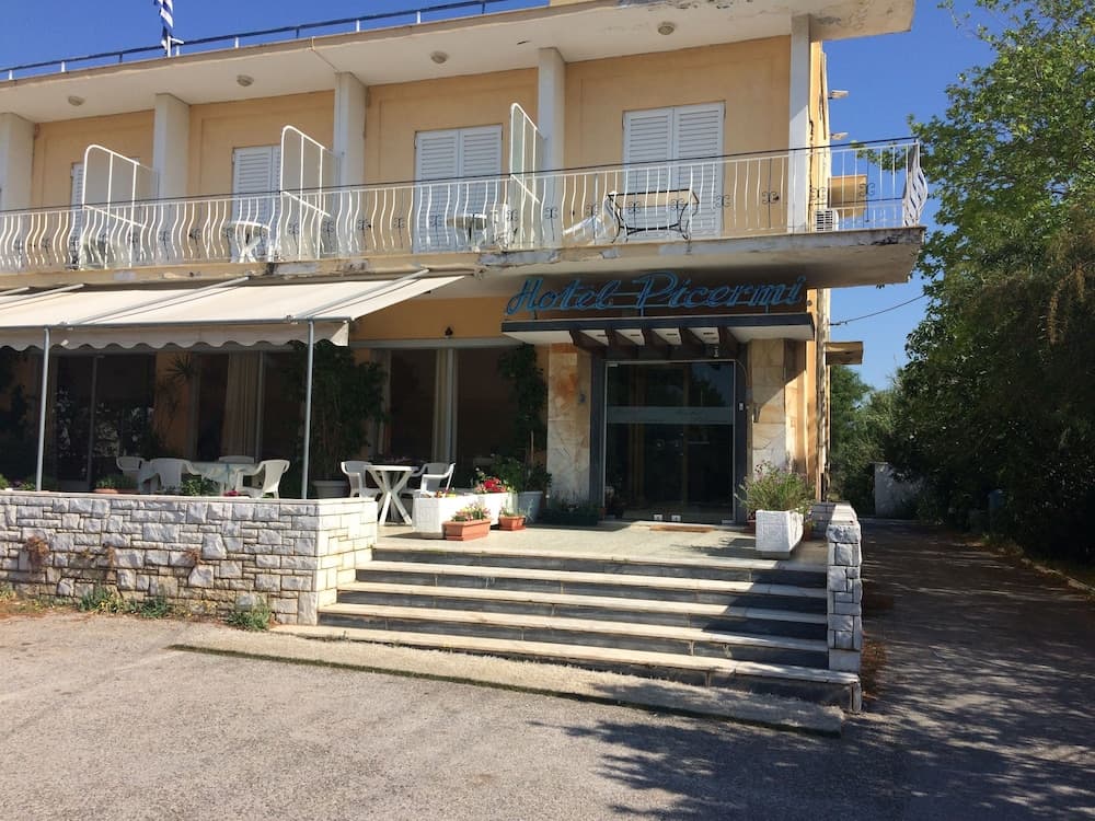 A serene hotel entrance features a yellow facade with white balconies, outdoor seating under an awning, and lush greenery, ideal for travelers seeking a tranquil Mediterranean retreat.