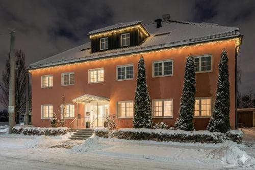 A cozy, warmly lit hotel adorned with festive lights stands invitingly in a snowy winter landscape, perfect for travelers seeking a serene and charming getaway.