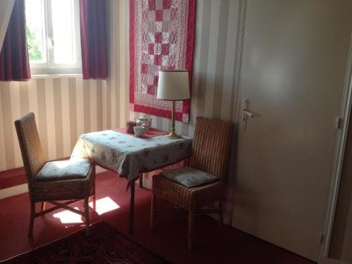 A cozy corner in a quaint guesthouse features a small table draped with a floral cloth, flanked by two cushioned chairs under soft sunlight streaming through a window, ideal for travelers seeking a peaceful retreat.