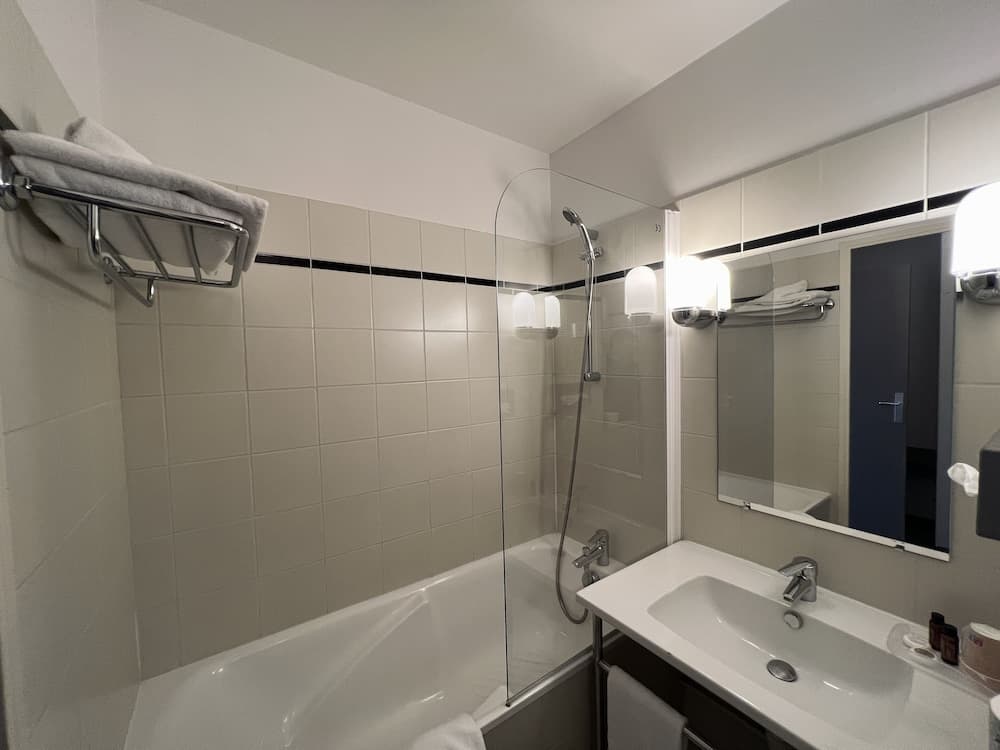A modern hotel bathroom features a sleek bathtub with a glass shower screen, a well-lit vanity mirror, and neatly arranged towels, ideal for travelers seeking comfort and cleanliness.