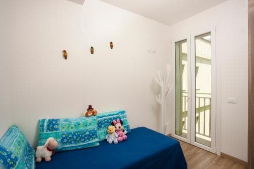 A cozy bedroom with a blue bed adorned with plush toys, white walls decorated with small ornaments, and a glass door leading to a balcony, ideal for a family-friendly vacation rental.
