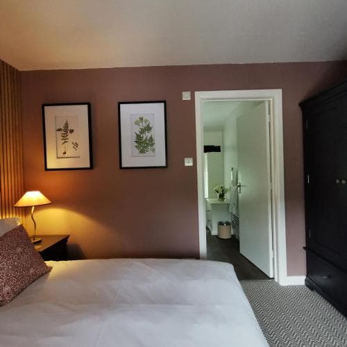 A cozy hotel room with a neatly made bed, framed botanical prints on the wall, a warmly lit bedside lamp, and an open door leading to a bathroom, ideal for travelers seeking comfort and relaxation.
