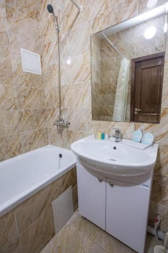 A cozy bathroom with beige marble tiles, a white bathtub, and a sink cabinet, featuring a large mirror and soft lighting, ideal for travelers seeking comfort in their accommodations.