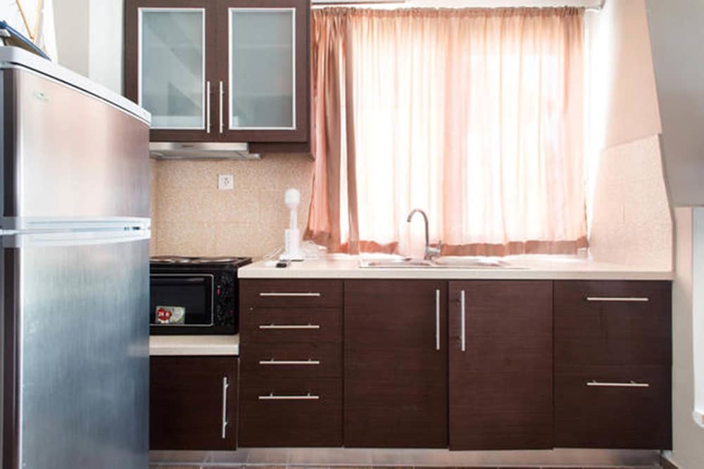 A cozy apartment kitchen featuring dark wooden cabinets, a stainless steel refrigerator, and a window with soft pink curtains, ideal for travelers seeking self-catering accommodations.