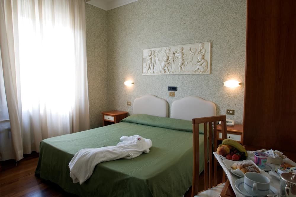 A cozy hotel room features twin beds with green linens, a decorative wall relief, soft natural light through sheer curtains, and a breakfast spread on a wooden table, ideal for travelers seeking comfort and convenience.