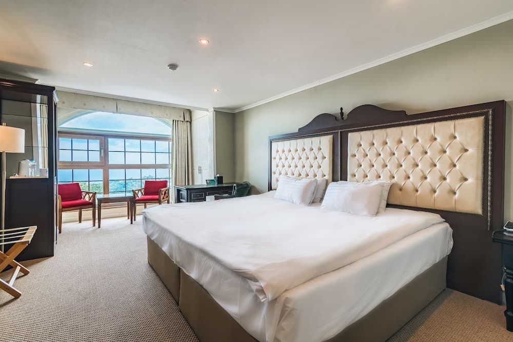 A serene hotel room with a plush bed, tufted headboard, and a scenic ocean view through large windows invites travelers seeking relaxation and comfort.