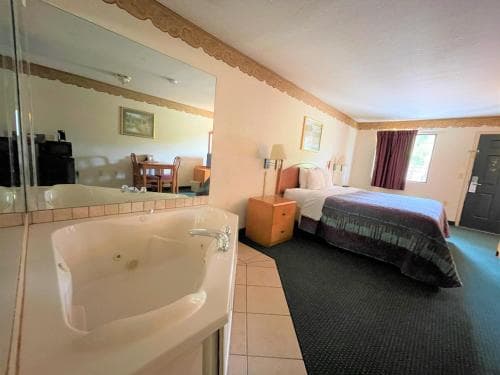A cozy motel room features a whirlpool tub in the foreground, a neatly made bed by the window, and a dining area reflected in a large mirror, ideal for travelers seeking relaxation and comfort.