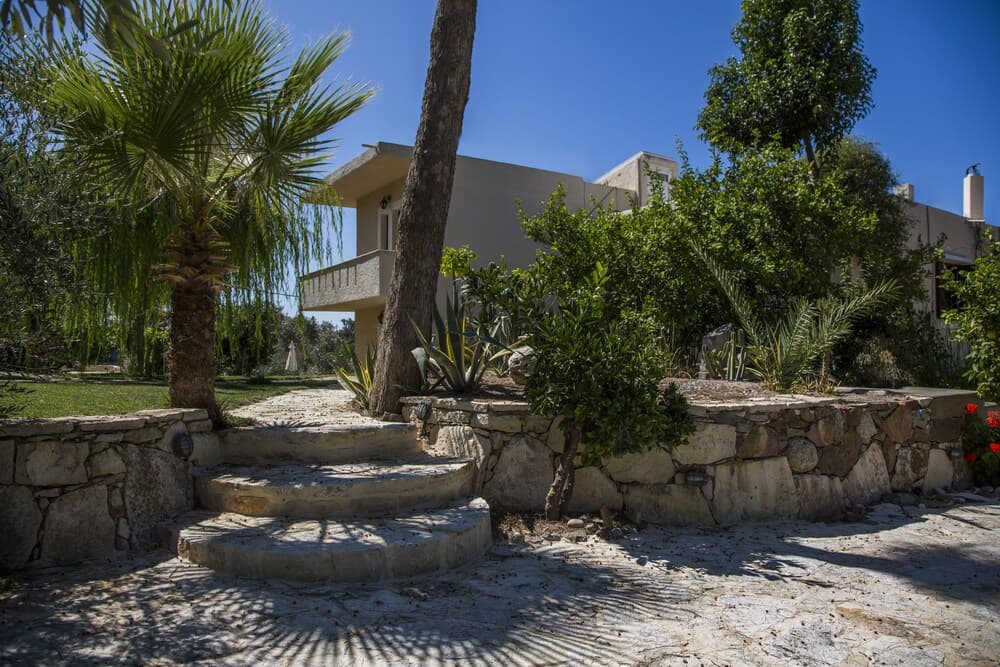 A serene Mediterranean villa surrounded by lush greenery, palm trees, and stone pathways invites travelers seeking a tranquil retreat in a picturesque setting.