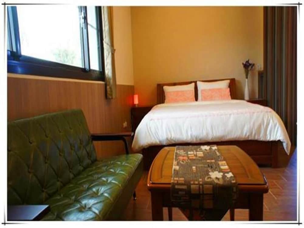 A cozy hotel room features warm wooden tones, a plush bed with pink accents, a green leather sofa, and a coffee table, creating an inviting atmosphere ideal for travelers seeking comfort and relaxation.
