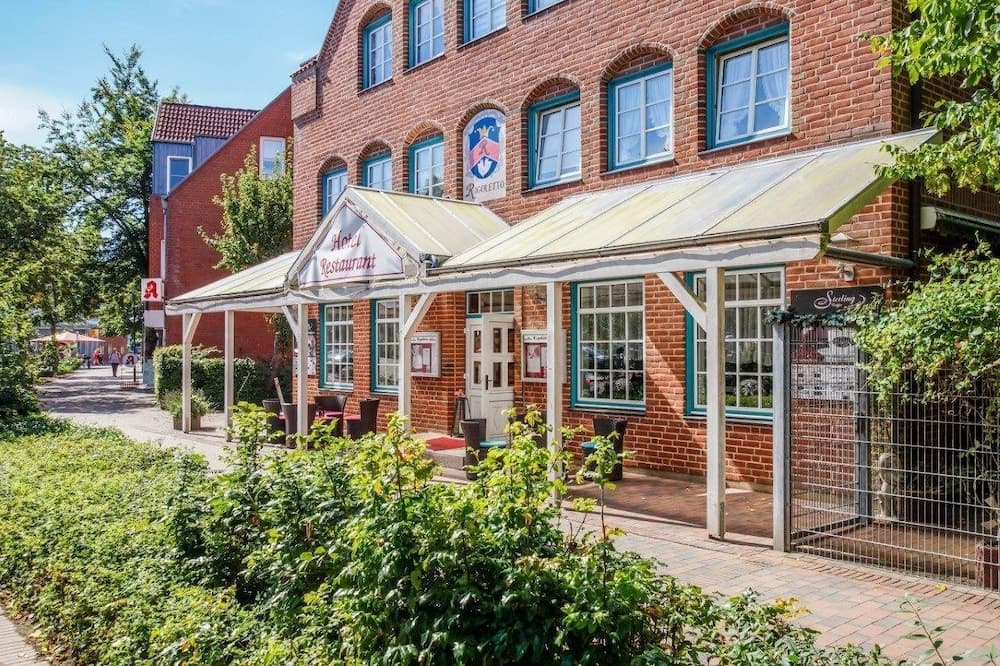A charming brick building with white-framed windows and a cozy outdoor seating area under a canopy invites travelers to enjoy a relaxing meal in a quaint European town setting.