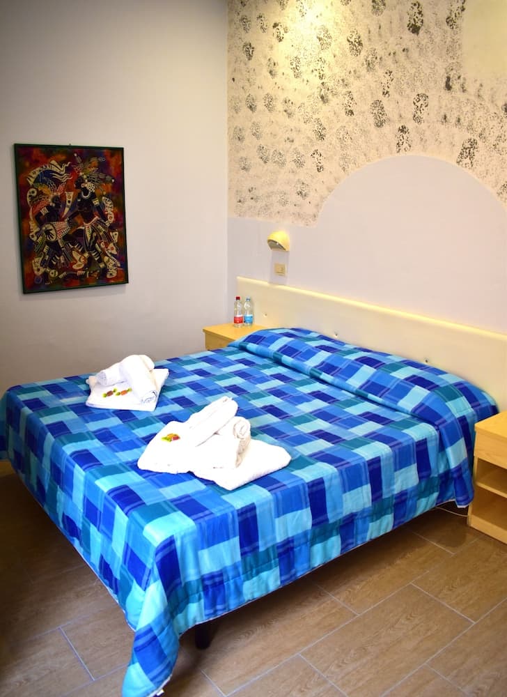 A cozy hotel room features a neatly made bed with blue plaid bedding, folded towels, a vibrant wall art piece, and a patterned wallpaper accent, ideal for travelers seeking comfort and charm.